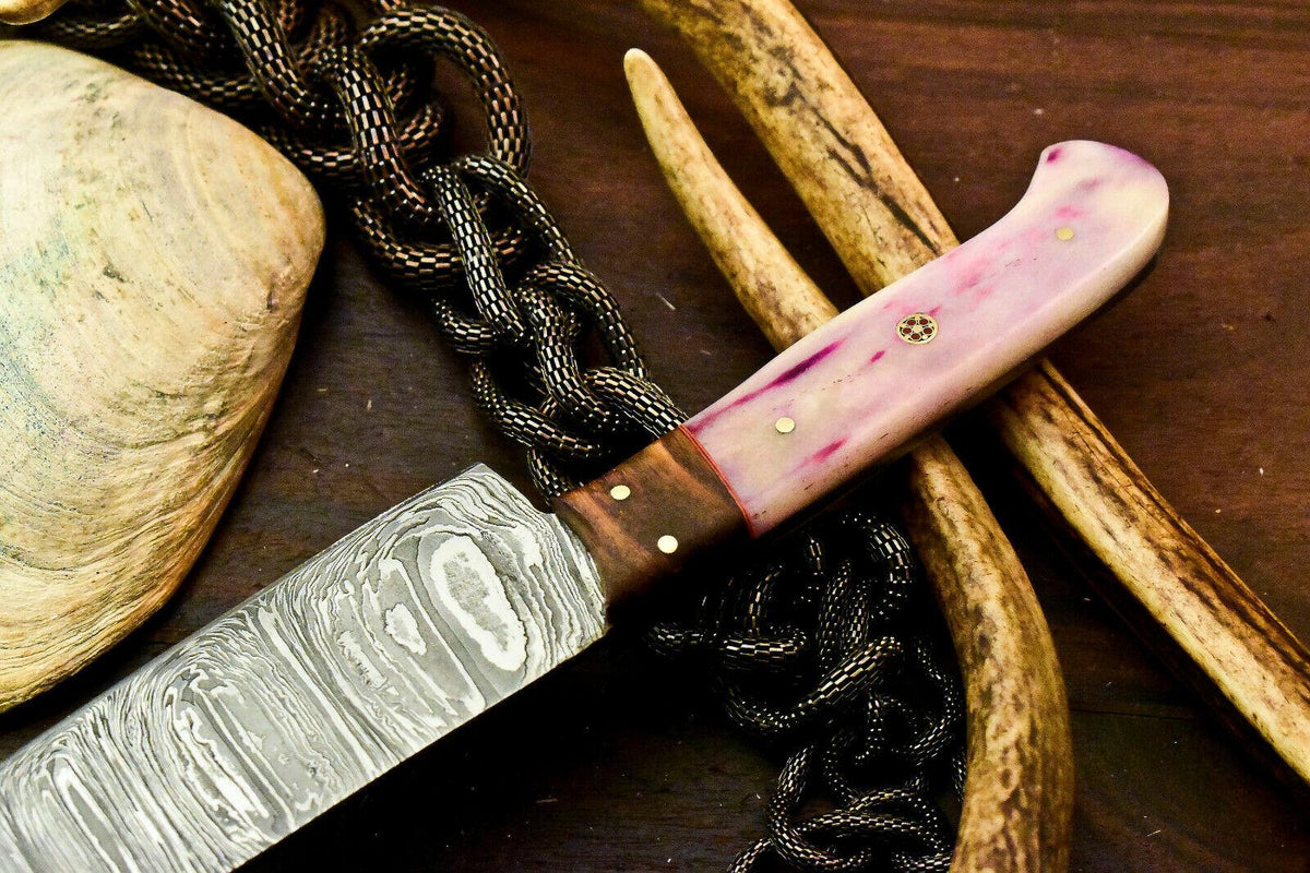 Close up of a custom Cosplay Swords kitchen knife showcasing a striking Damascus blade texture, a wooden bolster, and an ergonomic pink bone handle set against rustic antler and shell props.